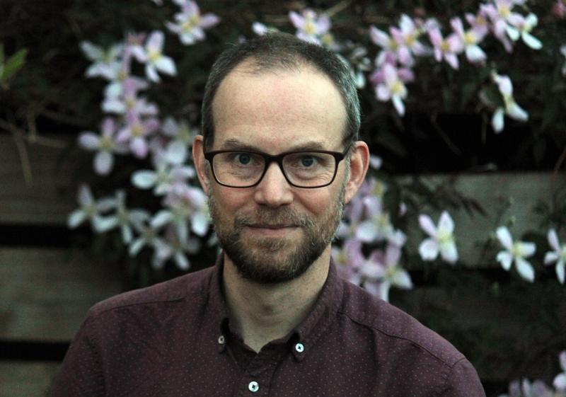 Head and shoulders shot of Bill Finnegan in front of a stone wall with flowers growing on it