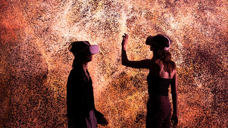 Two people wearing VR headsets in front of an explosion of orange light, which looks like millions of tiny dots