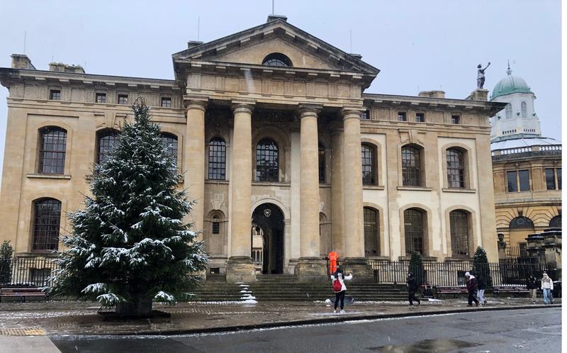 The photo shows the Clarendon Building with large columns and a snow-dusted Christmas tree in front. People in winter attire walk nearby. The Sheldonian Theatre's domed structure is visible in the background.